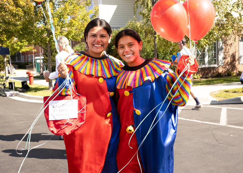 Two people in colorful clown costumes holding red balloons on a street.