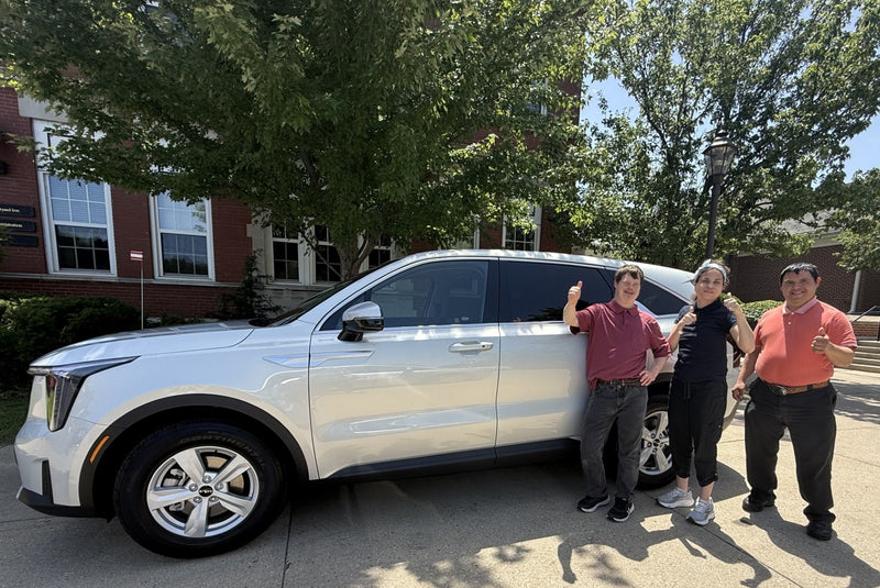 A white SUV parked outdoors on a driveway with three people standing beside it in front of trees and a brick building.