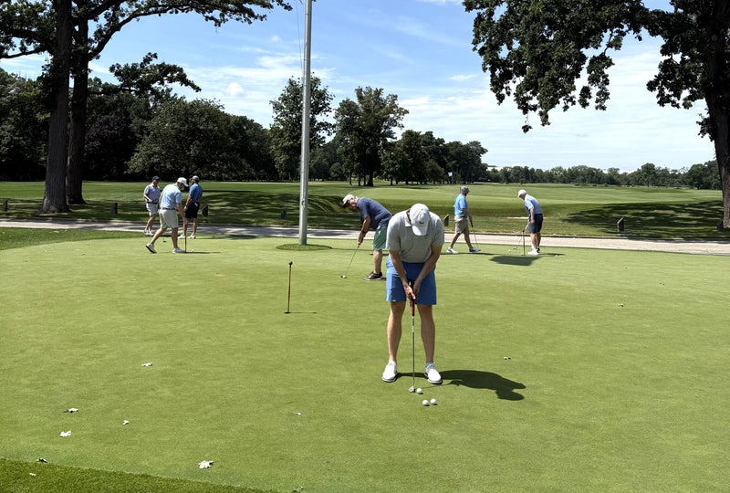 A man in a white hat, blue shorts, and white shirt is putting on a green golf course, with other players and trees in the background.