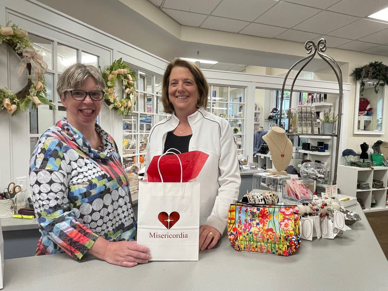 Two women standing behind a counter with a Misericordia bag and various items.