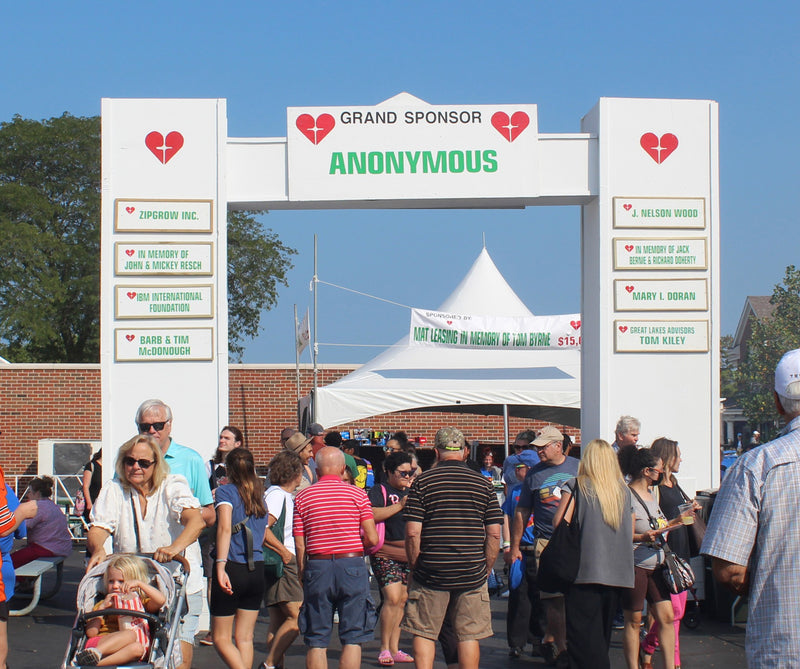 A crowd walking under a large white  sponsor arch at an outdoor event on a sunny day, with tents and people visible in the background.