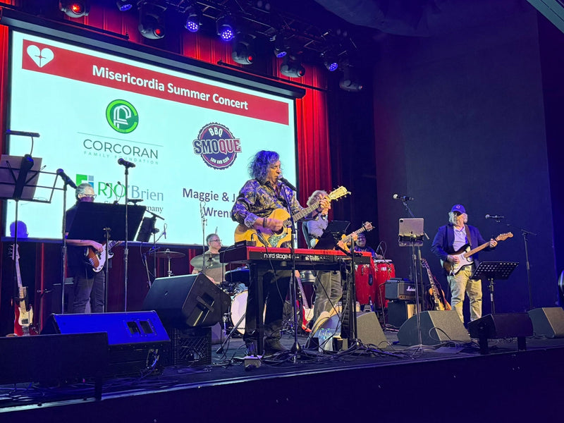 A live band performing on a stage in front of a large screen displaying 'Misercordia Summer Concert' and sponsor logos, including Corcoran and Smoque.
