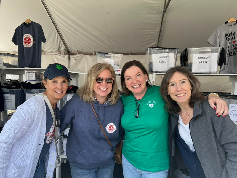 Four smiling women standing together under a white event tent, with T-shirts displayed on shelves behind them.